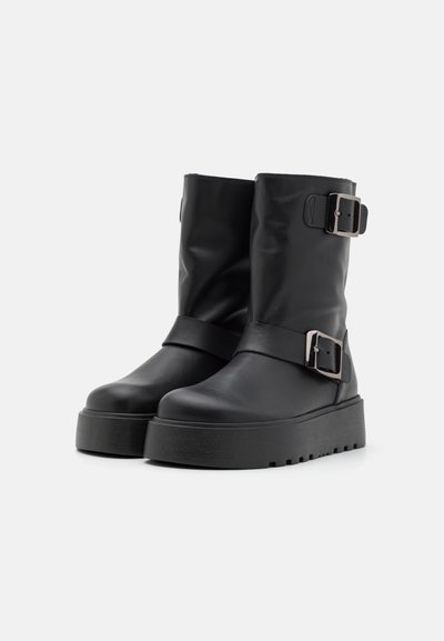 Bottes noires montantes en simili cuir lisse, dotées de deux boucles en métal ajustables et d'une semelle épaisse et texturée.