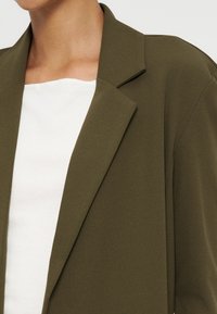Gros plan d’une personne portant un blazer vert olive par-dessus un haut blanc, montrant le col et le haut de la poitrine.