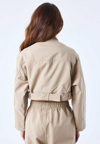 Beige cropped jacket van glad materiaal met een kraag, elastische taille en een riemdetail op de rug. Textuurpatroon zichtbaar op de stof.