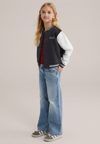 Črno-bele univerzitetne jakne s gumbi in logotipom, rdeča majica, svetlo modri uničeni jeans in sivi športni copati z črtami.