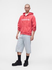 Homme debout portant un sweat à capuche rouge zippé avec le texte « SUNBURN », un short en jean clair, des chaussettes noires et des chaussures noires sur fond uni.