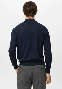 Granatowy sweter dzianinowy z długimi rękawami i kołnierzykiem, z prążkowanym obszyciem na dole i mankietach, noszony z szarymi, dopasowanymi spodniami. Widoczne minimalne detale.