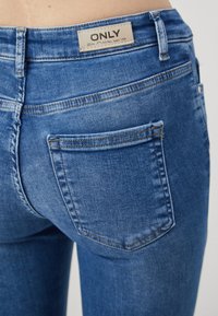 Vaqueros de mezclilla azul oscuro con una forma ajustada, que presentan bolsillos traseros y un detalle de costura clara. La etiqueta dice "ONLY" en la cintura.