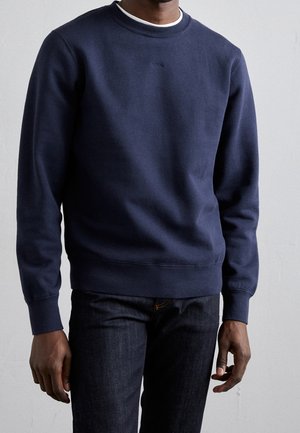 Hombre con sudadera azul marino con cuello redondo sobre camiseta blanca y vaqueros oscuros, de pie frente a un fondo gris liso.