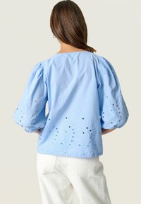 Blusa azzurro chiaro con maniche a palloncino, con motivi floreali ricamati e fori a occhio lungo l'orlo. Indossata con pantaloni bianchi texturizzati.