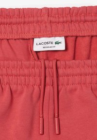 Pantalones jogger coral con cintura elástica, cordón y textura suave. Presenta una etiqueta "Lacoste" que indica un ajuste regular.