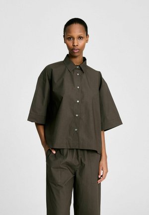Kvinde iført løs, mørkeolivengrøn skjorte med knapper og albuelange ærmer samt matchende højttaljede bukser, stående mod en ensfarvet lys baggrund.