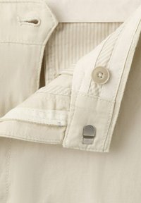 Gros plan sur la ceinture d'un pantalon beige montrant un bouton, une fermeture à crochet et barre, et une boutonnière cousue sur un tissu texturé.