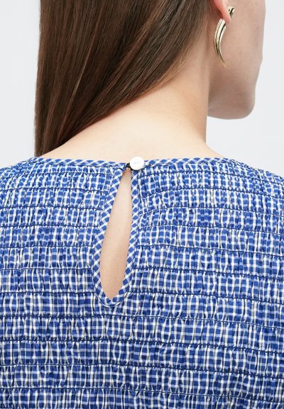 Femme portant un haut à carreaux bleu et blanc avec un tissu froncé et une petite ouverture en trou de serrure fermée par un bouton blanc au niveau de l'encolure arrière.