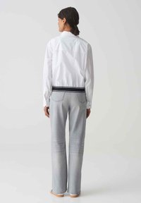 White shirt with a fitted back, featuring a contrasting black waistband. Paired with light gray flared jeans and simple footwear.