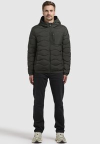 Dunkelgrüne Steppjacke mit einem gesteppten Muster, Frontreißverschluss und Kapuze; ausgestattet mit einer Brusttasche und schlankem Schnitt. Wird über dunklen Jeans und Schuhen getragen.