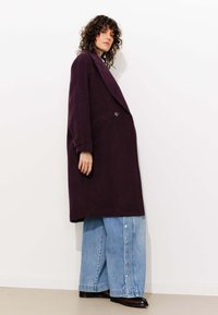 Manteau en laine violet surdimensionné avec un devant à double boutonnage, de grands revers et des poches latérales, associé à un jean en denim à jambe large avec des accents de boutons.