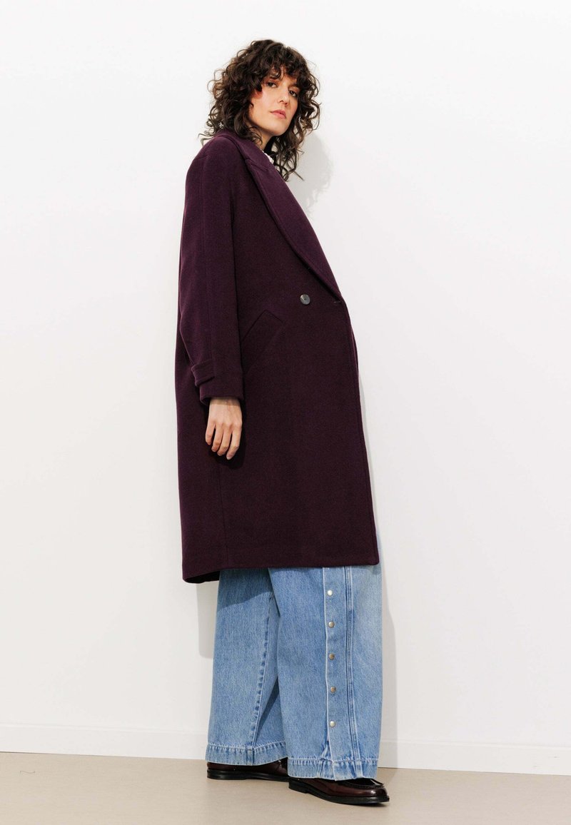 Manteau en laine violet surdimensionné avec un devant à double boutonnage, de grands revers et des poches latérales, associé à un jean en denim à jambe large avec des accents de boutons.