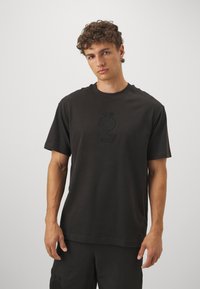 Svart t-shirt med kort ärm i bomull, med en subtil präglad logotyp på bröstet. Klassisk rundhalsad design, avslappnad passform.