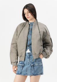 Mlada ženska nosi prevelik sivi bomber jakno preko modrega denim topa in ujemajoče krilo z velikimi sprednjimi žepi, stoji ob beli ozadju.