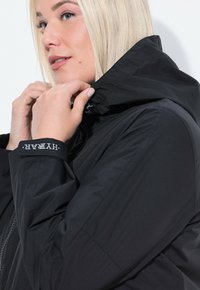 Una donna con i capelli biondi aggiusta il colletto di un giubbino nero con dettagli agli polsini marchiati "HYBAR" su uno sfondo bianco.