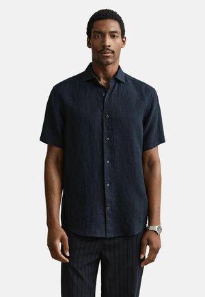 Man draagt een marineblauw kort mouw overhemd met knopen en donkere gestreepte broek, staand tegen een effen witte achtergrond.