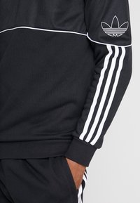 Primer plano de una persona que lleva un chándal negro de Adidas con rayas blancas en la manga y los pantalones, con la mano descansando en el bolsillo.