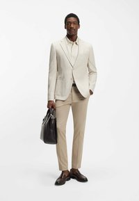 Beige gestructureerde blazer over een lichtbeige polo, gecombineerd met slanke beige broek en bruine leren schoenen, met een donkerbruine tas in de hand.