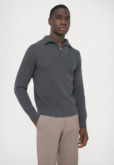 Camisola cinzenta com gola de meio fecho éclair, textura canelada e mangas compridas; usada com calças castanho claro. Design simples, sem padrões.