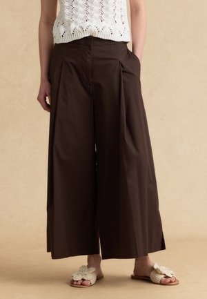 Donna che indossa pantaloni ampi marrone scuro, top lavorato a maglia bianco senza maniche e sandali beige aperti con fiocchi decorativi.