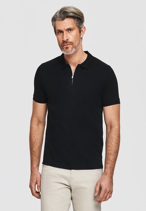 Mężczyzna z siwymi włosami i brodą, ubrany w czarną polo z krótkim rękawem i zamkiem z przodu, stojący na jasnoszarym tle.