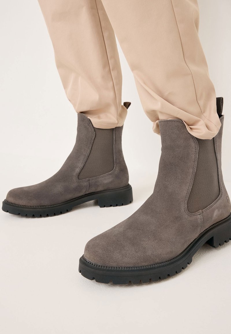 Bottes Chelsea en daim taupe avec semelles épaisses noires portées avec un pantalon beige, mettant en valeur les panneaux élastiques sur les côtés et les languettes à l'arrière.