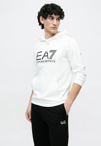 Mladý muž s kučeravými vlasmi, oblečený v bielej mikine EA7 Emporio Armani a čiernych nohaviciach s logom EA7, ruky v kapse, pozerá sa do strán.