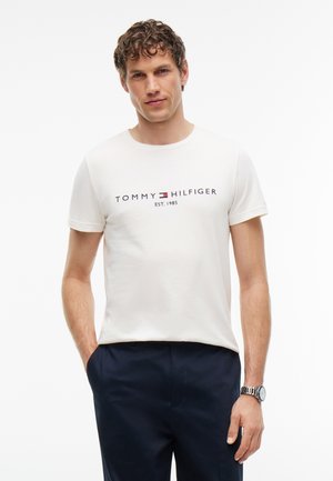 Homme aux cheveux bouclés portant un t-shirt blanc Tommy Hilfiger et un pantalon foncé, debout les mains dans les poches devant un fond uni.