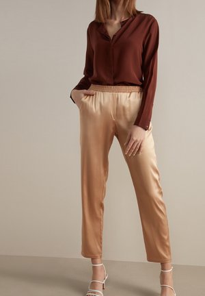 Kvinde iført en brun bluse med lange ærmer, beige satinbukser med elastisk talje og hvide højhælede sandaler med stropper, står mod en ensfarvet baggrund.
