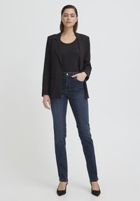 Femme debout portant un blazer noir, un haut noir, un jean bleu foncé et des talons noirs, posant avec une main dans la poche du jean sur un fond uni.