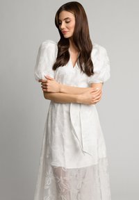 Biała sukienka z bufkowymi rękawami, zdobiona kwiecistym wzorem i przezroczystą warstwą. Długa, z przewiązanym w talii paskiem. Gładka faktura materiału.