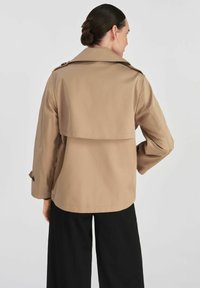 Beżowy trencz o dopasowanej sylwetce i rozkloszowanej tylniej części. Posiada długie rękawy, detale z guzikami i gładki, lekki materiał.