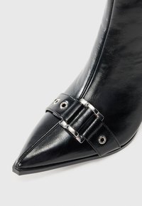 Scarpa nera in ecopelle a punta con un dettaglio decorativo a fibbia e accenti in metallo argentato. Texture liscia con una finitura elegante.
