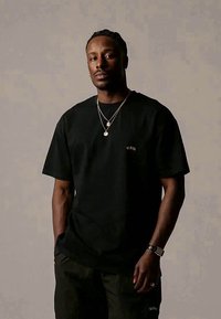 Black cotton t-shirt with a round neck, featuring a small embroidered logo. Model wears slim-fit black pants and layered silver necklaces.