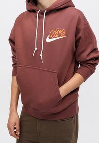 Personne portant un sweat à capuche marron avec des cordons blancs, un motif orange et blanc sur la poitrine, et une main dans la poche avant.