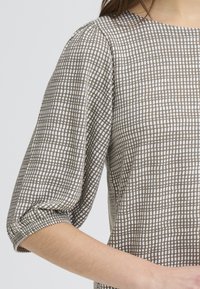 Haut à carreaux marron et blanc avec un col rond et des manches bouffantes trois-quarts, en tissu doux avec une finition texturée.