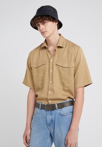 Short-sleeve khaki shirt with two chest pockets, beige jeans, and a black bucket hat. Soft texture, button-down design, casual fit.