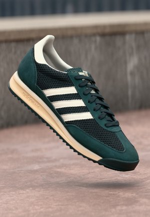 Basket vert foncé avec des bandes blanches, semelle beige et lacets noirs, montrée en l'air au-dessus d'une surface en béton.