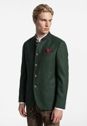 Junger Mann trägt eine dunkelgrüne traditionelle Jacke mit silbernen Knöpfen, weißem Hemd, rotem Einstecktuch und brauner bestickter Hose und schaut zur Seite.