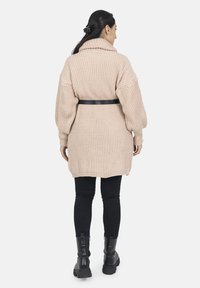 Cárdigan de punto beige de gran tamaño con un cuello alto, cintura ceñida y textura acanalada, combinado con jeans ajustados negros y botas gruesas.