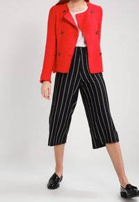 Veste texturée rouge avec de grands boutons noirs, assortie à des culottes à rayures noires et blanches. Des chaussures noires complètent la tenue.