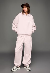 Svetloružová oversized mikina a zodpovedajúce tepláky, oboje s bielym textom, v kombinácii so striebornými a čiernymi teniskami, na sivom pozadí.