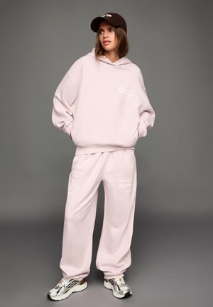 Svetloružová oversized mikina a zodpovedajúce tepláky, oboje s bielym textom, v kombinácii so striebornými a čiernymi teniskami, na sivom pozadí.