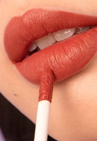 Matte liquid lipstick in a rich terracotta shade applied to full lips. A precision applicator is poised above the lips for application.