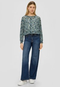 Bluse mit floralem Muster in Blau und Grün; lange Ärmel und runder Ausschnitt. Kombiniert mit weit geschnittenen, dunklen Jeans und weißen Sneakers.