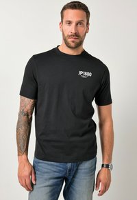 Czarna bawełniana koszulka z krótkim rękawem, z białym logo "JP1880" na lewej piersi. Noszona z niebieskimi dżinsami i zegarkiem na nadgarstku.