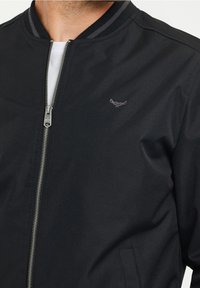 Veste bomber noire avec une fermeture éclair à l'avant, un col côtelé avec des rayures grises, et un petit logo brodé sur la poitrine gauche.