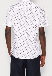 Chemise à manches courtes avec un fond blanc et un imprimé floral rouge, dotée d'un col boutonné et d'un ourlet droit. Texture de tissu lisse.