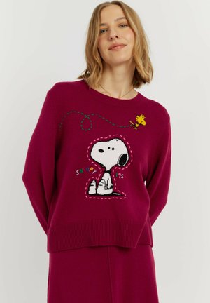 Vrouw draagt een paarse trui met een cartoonontwerp van Snoopy en Woodstock, gecombineerd met een bijpassende paarse broek tegen een effen achtergrond.